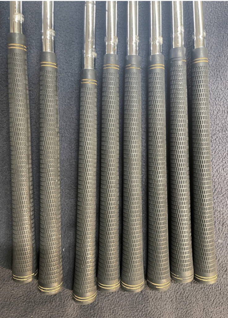 Set of black textured golf club grips on chrome shafts laid out on a dark surface, ready for inspection and conditioning